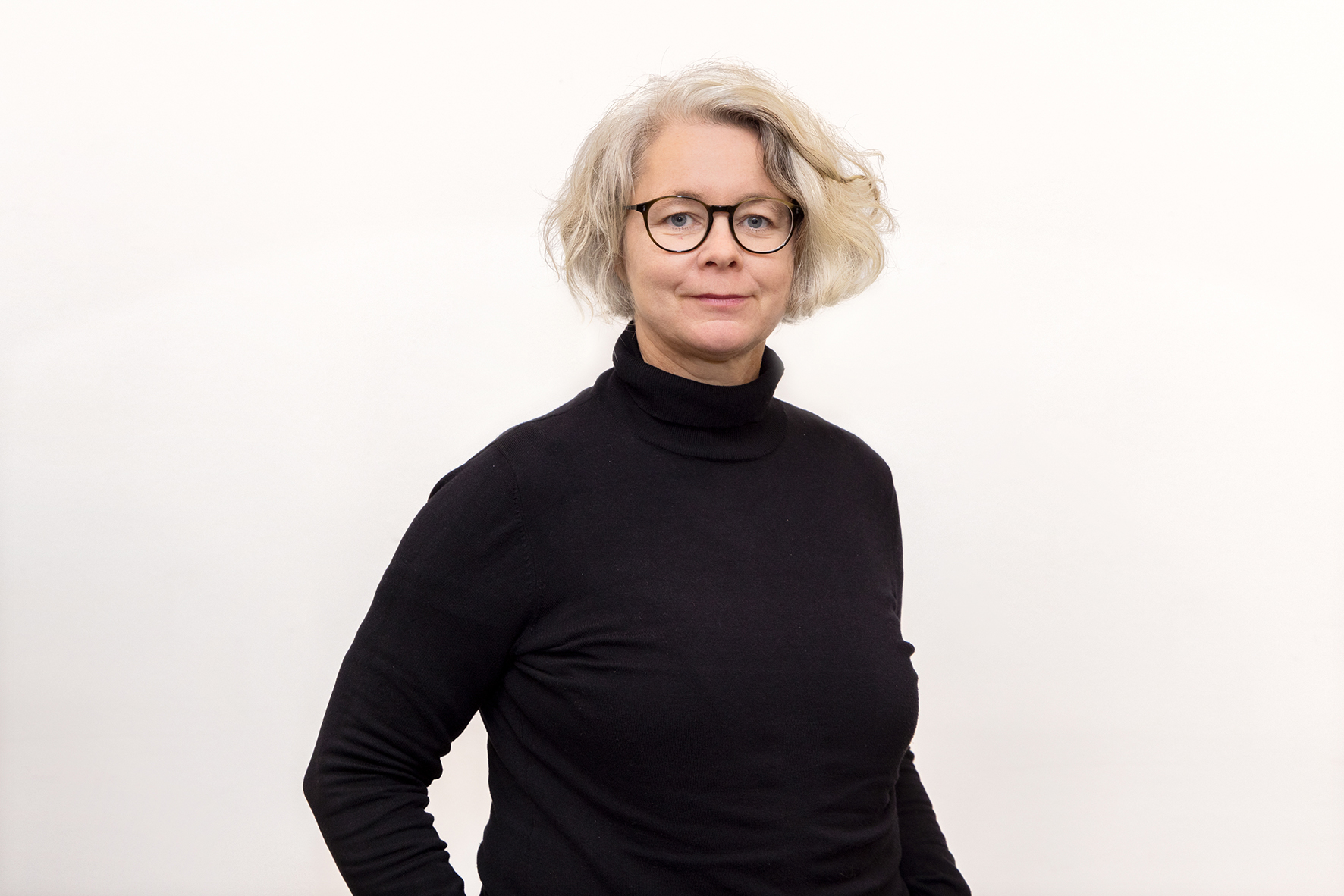 Verena schaut mit einem  Lächeln in die Kamera. Sie steht vor einer weißen Wand. Verena hat einen schwarzen Rollkragenpulli an. Sie trägt eine schwarze rundliche Brille.